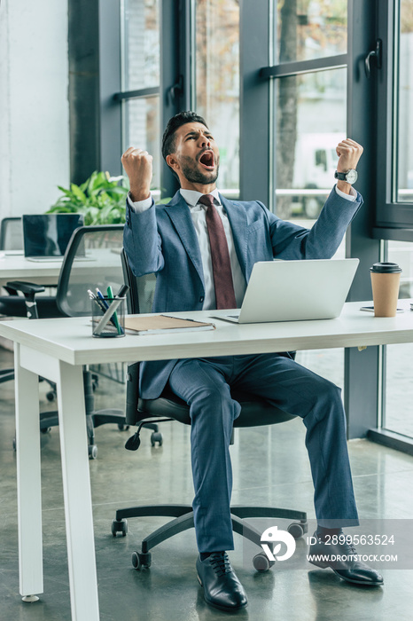 happy businessman screaming and showing winner gesture while sitting at workplace