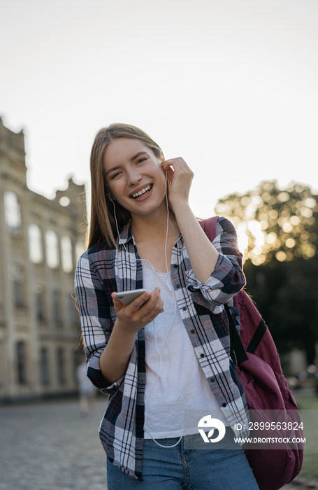 Student with backpack walking to university, listening music, laughing outdoors. Young cheerful woman using mobile application for smartphone, chatting, communication, online shopping, ordering food