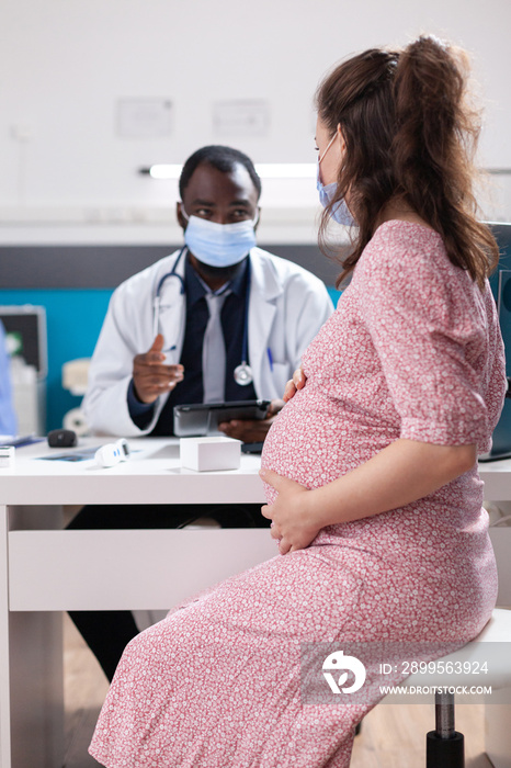 Physician doing medical consultation with pregnant woman, wearing face masks. Woman expecting child and receiving medical advice from doctor at office desk during covid 19 pandemic