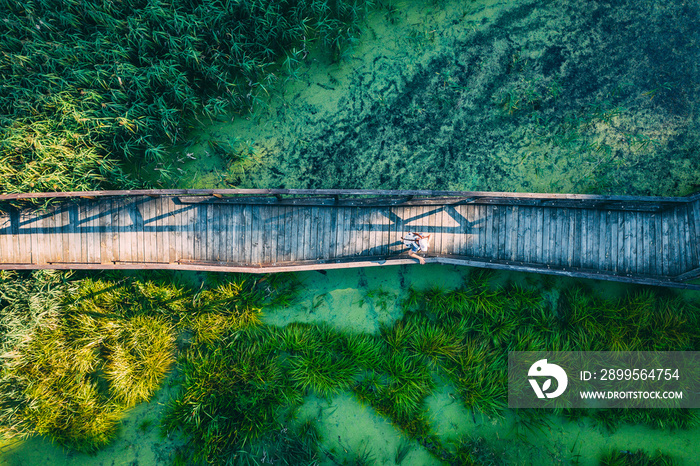 Aerial top view of woman traveller on wooden bridge pathway over marshy river with vegetation thickets, summer travel concept