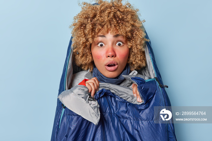 Shocked female hiker stares with omg expression cannot believe own eyes has red frozen cheeks poses in sleeping bag spends leisure time in nature isolated over blue background has adventure trip