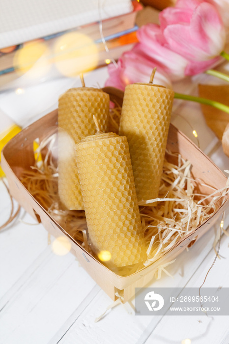 Candles from the honeycomb lie in a basket near pink lulips, books and fairylights on the background of a wooden table.