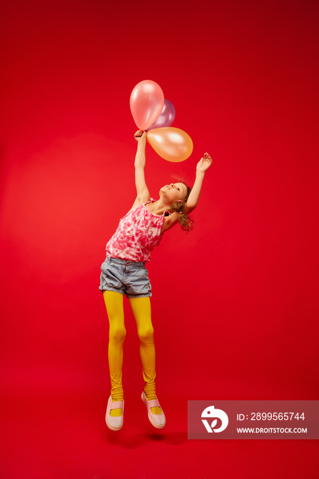 Little cute girl, child with braids emotionally posing, playing, jumping with air balloons on bright red studio background. Concept of childhood, emotions, fun, fashion, lifestyle, facial expression