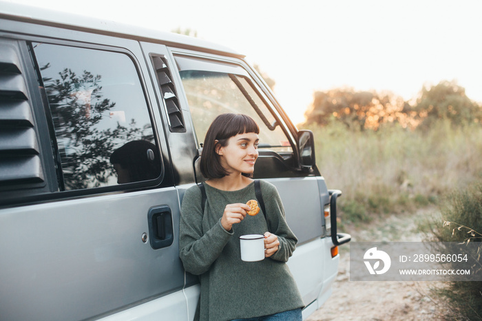 Beautiful young girl travels around world in camping van, looks to side while enjoying cup of hot coffee or tea from hiking cup or mug, smiles, concept millennial adventures and hipster lifestyle