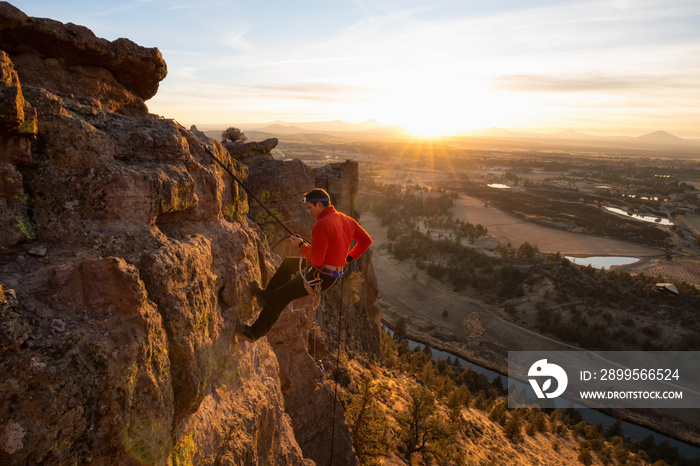 Adventurous man is rappeling down a cliff during a bright and vibrant sunny sunset. Taken in Smith Rock, Oregon, North America.