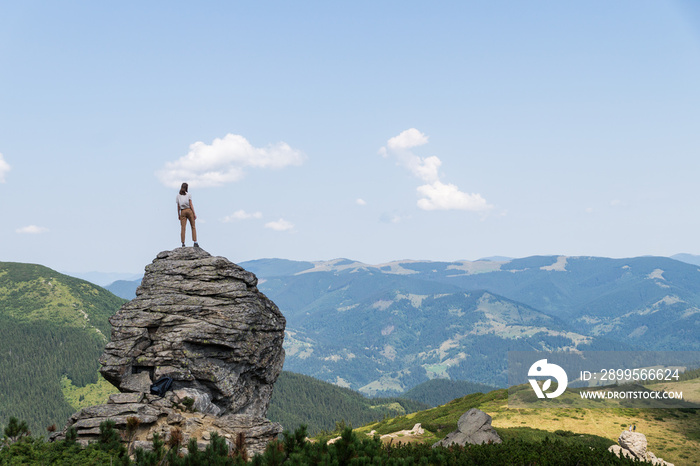 Young female tourist stands on top of the rock in rural mountain area and looks at beautiful panoramic scenery of the ukrainian carpathian mountains