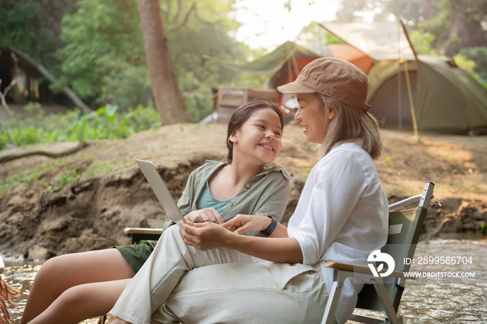 Pleased happy mother and daughter reading a book and using laptop while relaxing on the deck chairs in the river, sit near a camp and tent, drink coffee in a pine forest. Camping, recreation, hiking.