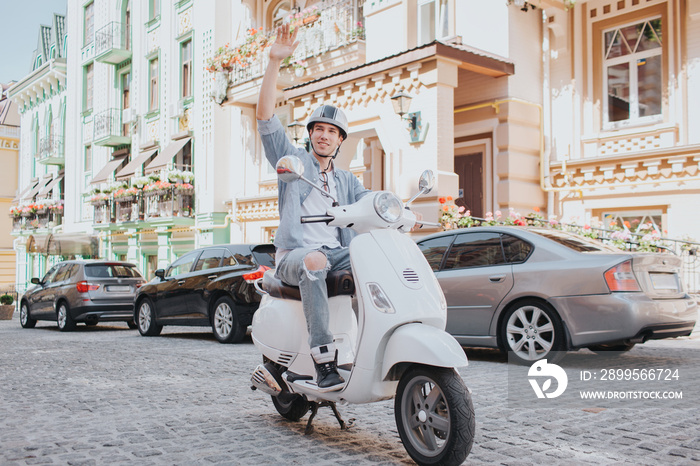 Man in casual clothes is riding on motorcycle. He is wearing helmet on head. Guy is holding fist in air and looking to the side. He is riding on road.