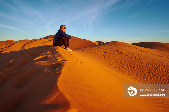 Blonde tourist girl watching sunrise in the desert sitting on a sand dune in Sahara Morocco Africa