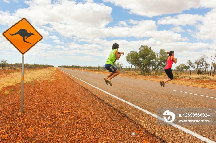 Funny couple of tourists pretend jumping like kangaroos by road sign on traditional empty highway in Northern Territory, Outback Australia