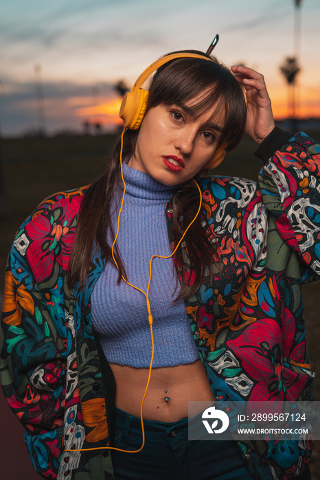 Young millennial woman listening to music with yellow headphone in an urban park during colorful sunset