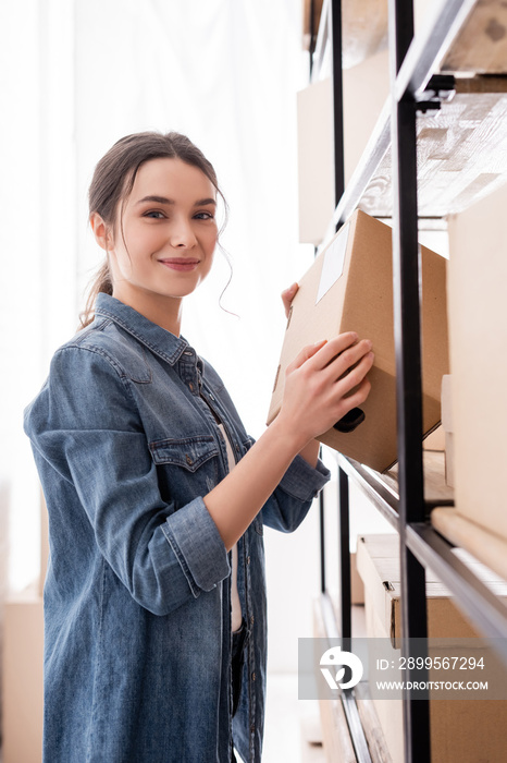 Smiling seller holding box and looking at camera near rack in online web store.