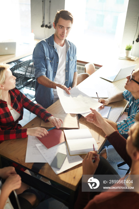 Group of creative business professionals discussing ideas while collaborating on startup project during meeting in modern office, focus on handsome young man handing out documents