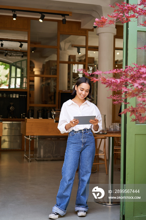 Young asian woman with tablet, standing in front of cafe entrance, inviting guests and customers, smiling broadly at camera