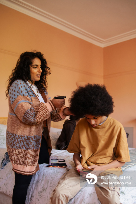 Mid-sized mother helping her son with his hair while he plays on his phone