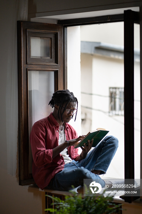 Pensive African American young man in eyeglasses reading sad book sitting on sill at home. Concentrated hipster male get knowledge from literature. Black student guy intently study difficult textbook.