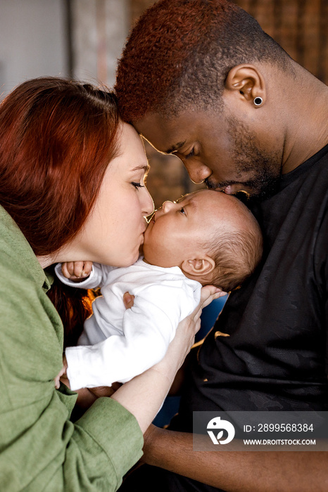 Happy multiracial mixed family gently holding newborn baby. Red haired caucasian mom looking at her child, Afro American father lovingly kissing cute baby in head