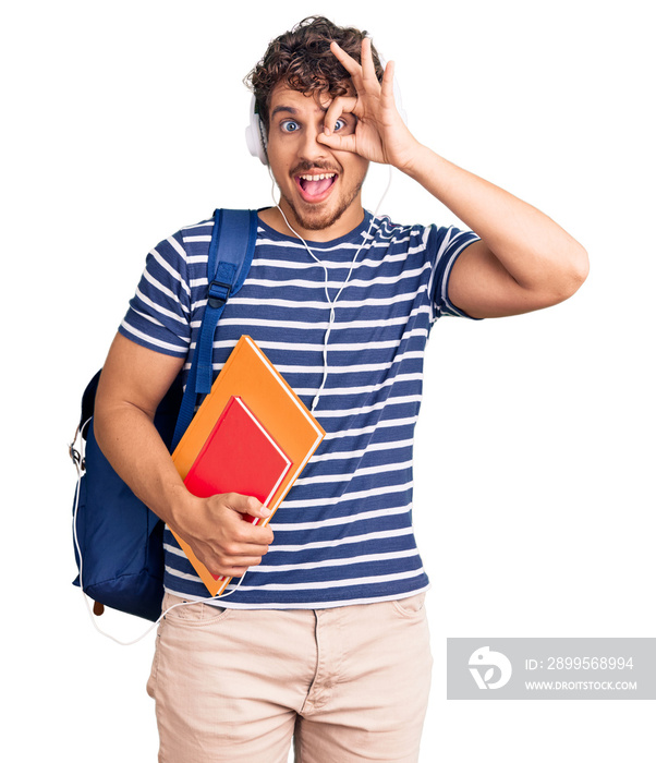 Young handsome man with curly hair holding student backpack and books smiling happy doing ok sign with hand on eye looking through fingers