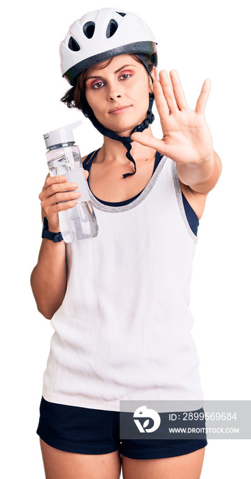 Beautiful young woman with short hair wearing bike helmet and holding water bottle with open hand doing stop sign with serious and confident expression, defense gesture