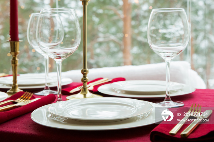Beautiful table setting with classic white porcelain, golden cutlery and candles. Big window with winter landscape, pine tree forest covered with snow. Christmas celebration