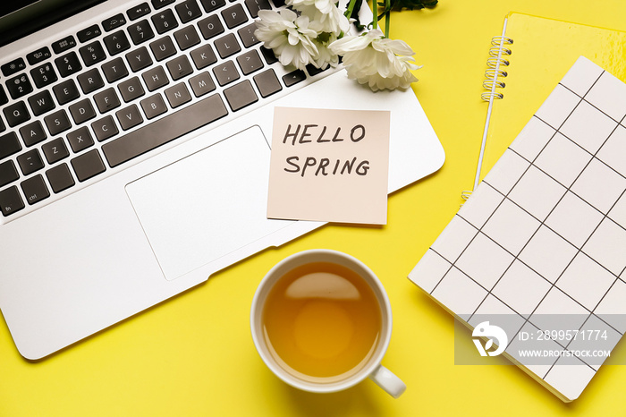 Paper sheet with text HELLO SPRING, laptop and cup of tea on color background