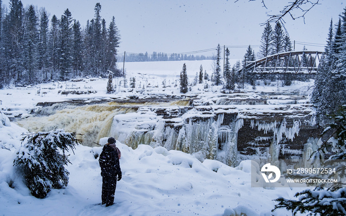 Woman wearing winter clothes standing in snow in front of a frozen waterfall in winter near Saguenay, Quebec (Canada)