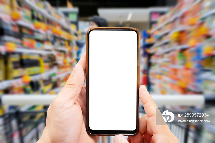 A person’s hand holds a mobile phone, with a shopping basket on the screen. In the background, a pretty woman selects digital products, in a blur. Mock up.