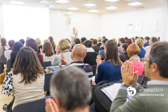 Life coaching symposium. Female speaker giving interactive motivational speech at entrepreneurship workshop. Audience in conference hall. Rear view of unrecognized participant.