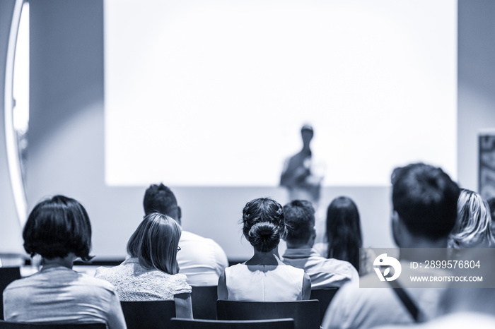 Speaker giving a talk in conference hall at business event. Focus on unrecognizable people in audience. Business and Entrepreneurship concept. Blue toned greyscale image.