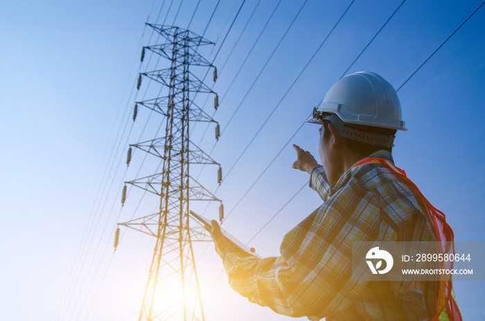 Electrical engineer holding and using a digital tablet, Engineer pointing at high voltage power pylon against sunset background