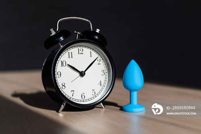 Blue latex anal plug and alarm clock on a wooden table. The time of carnal pleasures.