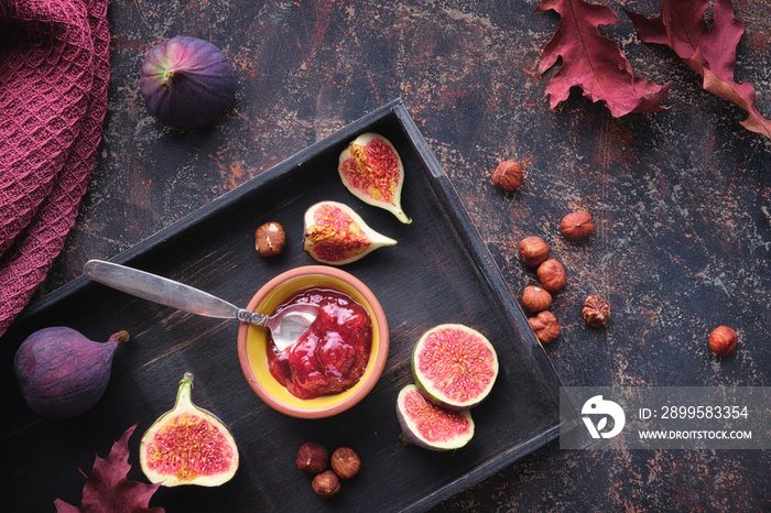 Autumntime background with fig jam, spoon, nuts and fresh halved fig fruits on black wooden tray. Dry red oak, ginkgo leaves on dark brown textured background. Seasonal Autumn, Fall deco arrangement.