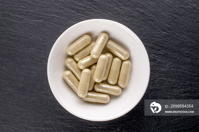 Several medical pills in a white ceramic bowl on a slate stone, close-up, top view.