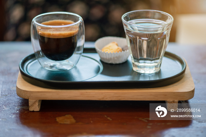 Hot espresso coffee in a clear glass, placed on a metal tray and wooden layer beside the coffee cup with a bear shaped cookie . All laid on dark brown wooden tables in retro coffee shop.