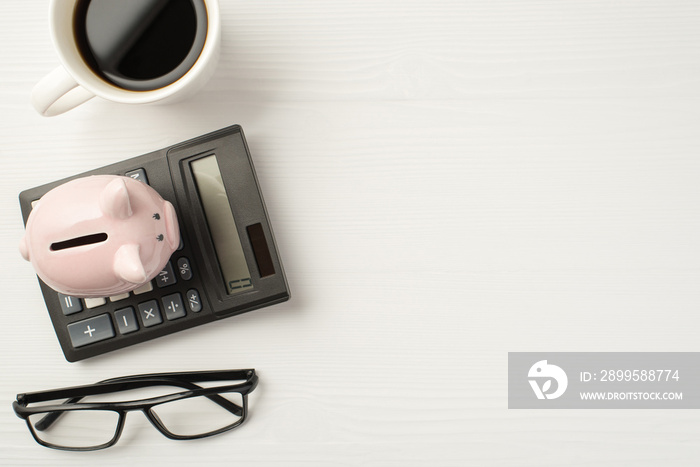 Flatlay top view photo of calculator with piggybank cup of espresso and eyeglasses isolated white wooden backdrop with empty space