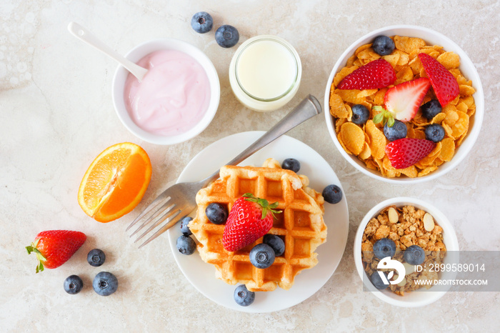 Breakfast food table scene. Fruits, cereal, waffles, yogurt and milk. Top view over a bright stone background.