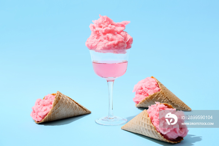 Glass with tasty cotton candy cocktail and waffle cones on color background