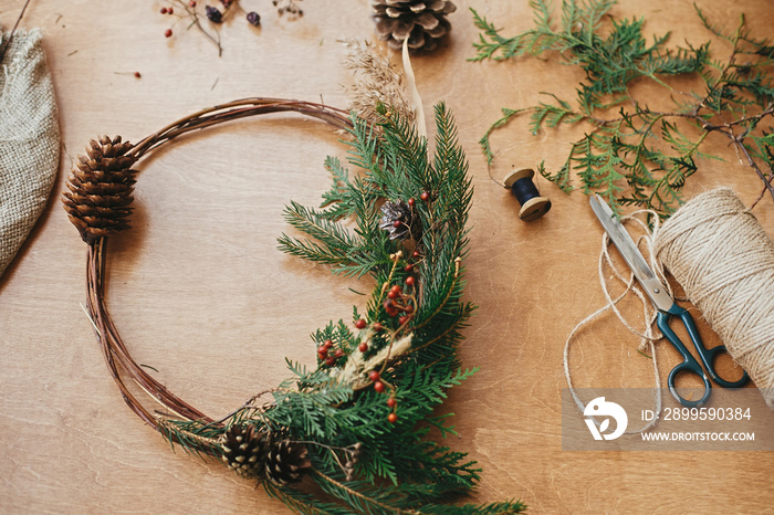 Rustic Christmas wreath. Creative christmas wreath with fir branches, berries, pine cones and herbs with scissors and thread on rural wooden table. Happy holidays. Workshop