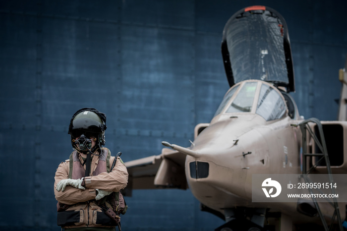 Fighter Pilot wearing helmet and visor standing next to fighter jet against a hangar door. Sand camouflage flying suit and jet. Visor down and oxygen mask on