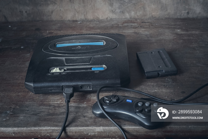 An old game console in black, in the dust on a wooden table, next to the joystick and cartridge.