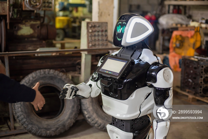 Humanoid robot on wheels with a monitor on his chest, shaking hands with a man. The old cluttered garage.