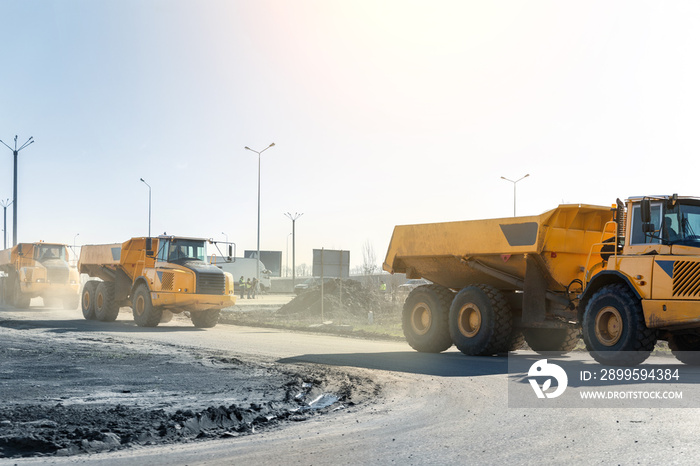 Many big articulated heavy industrial yellow dumper trucks driving on new highway road construction site on sunny day with blue sky background. Construction equipment machinery working on open pit