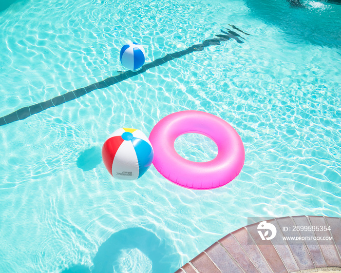Bright pink float and beach balls in blue swimming pool, floating in refreshing swimming pool with waves reflecting in summer sun. Active vacation background. Lifesaver for kid. Sunny day at the pool