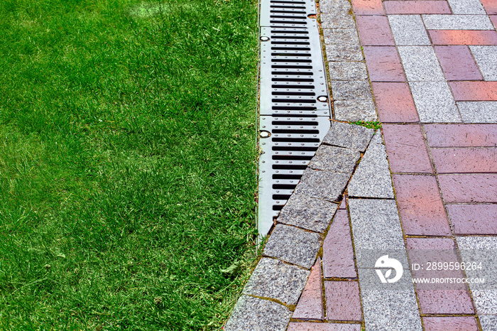 drainage grate bolted to storm drain at corner of pedestrian pavement walk way made of stone brick tiles gray and red pattern in backyard with copy space on green grass on park, nobody.