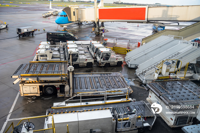 Luggage carts and other equipment for loading and unloading aircrafts on the tarmac of an airport. An airplane connected to an air bridge is in background. Schiphol airport, The Netherlands.