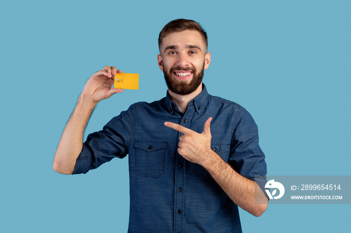 Joyful millennial man pointing at credit card on blue studio background. Electronic money concept