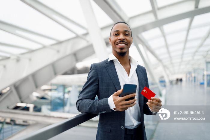 Young Black Businessman Using Smartphone And Credit Card While Standing In Airport