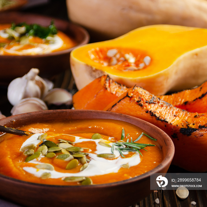 Homemade rustic pumpkin soup with seeds in clay dish on wooden table with bread spices and greens as