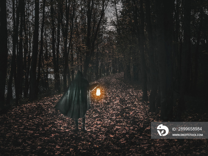 Man in motion in black brilliant hooded cloak holding the glowing lantern on the dark forest