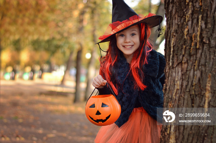 Halloween trick or treat.Happy girl wearing a witchs hat, with a pumpkin handbag, peeps out from be